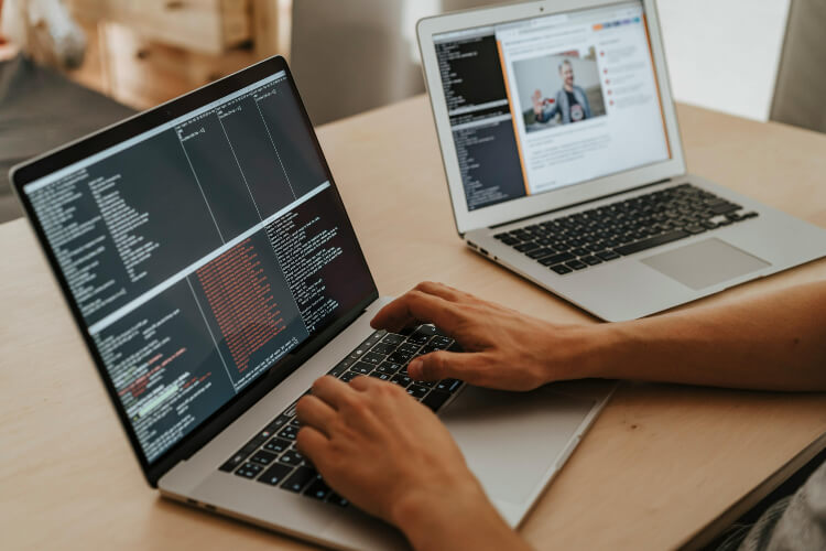 person coding on one laptop, with another laptop left with tabs open on the other side of the desk.