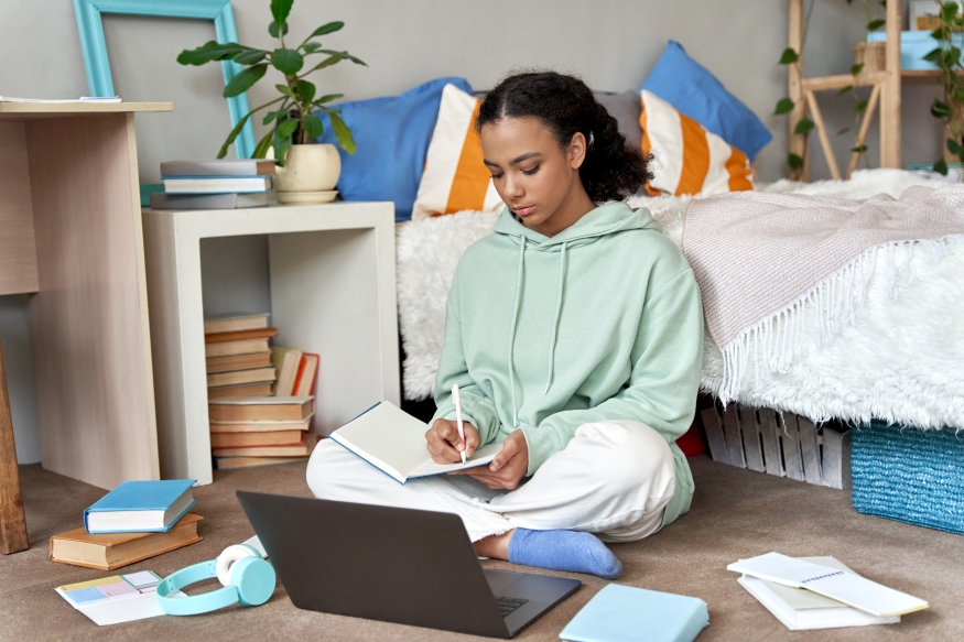 A young woman sitting next to her bed with her laptop and paper surrounding her.