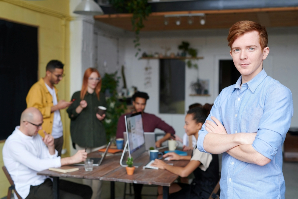 A young person with arms folded looking at the camera, in front of a table of work colleagues gathered around a laptop.